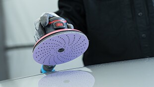 Main gantée avec ponceuse orbitale 3M et un disque de ponçage perforé sur surface lisse.
