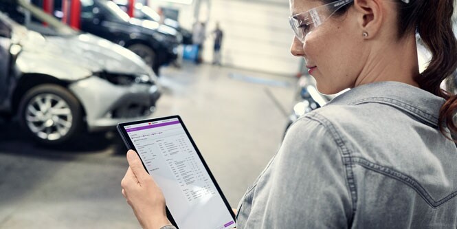 Une femme, debout dans un atelier de carrosserie, consulte l'écran d'une tablette.
